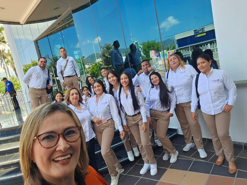 En la imagen se ve un grupo numeroso de personas posando en unas escaleras a la entrada de un edificio moderno con fachada de vidrio.
En primer plano aparece una mujer con gafas y blusa naranja, esa mujer es Zonia Vergara, está sonriendo a la cámara tipo selfie. Al fondo, sobre los escalones, hay alrededor de 15 personas más, los cuales son sus estudiantes, la mayoría mujeres jóvenes, todas muy arregladas, usando un uniforme similar: camisa blanca de botones y pantalón beige, muchos con tenis blancos y algunos con morral al hombro.
Todos se ven sonrientes y relajados, como si estuvieran celebrando o registrando un momento especial en grupo, posiblemente de estudio, trabajo o una visita institucional. El reflejo en los vidrios muestra el cielo azul, árboles y parte de la ciudad, lo que da sensación de día soleado y ambiente agradable.